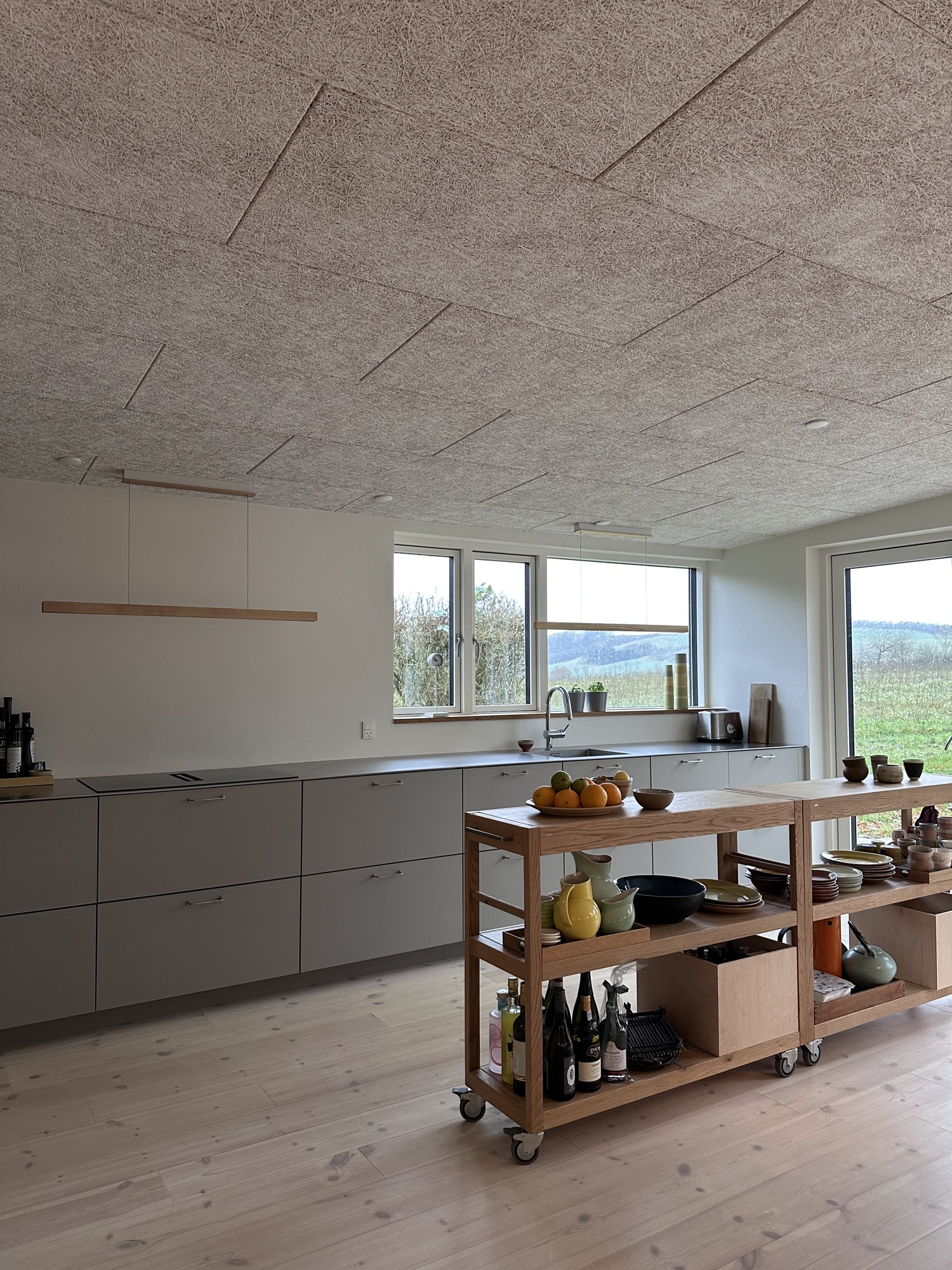 Woodfront-køkken på Ringvej med greige fronter, træbetonloft og vinduesbånd mod markerne