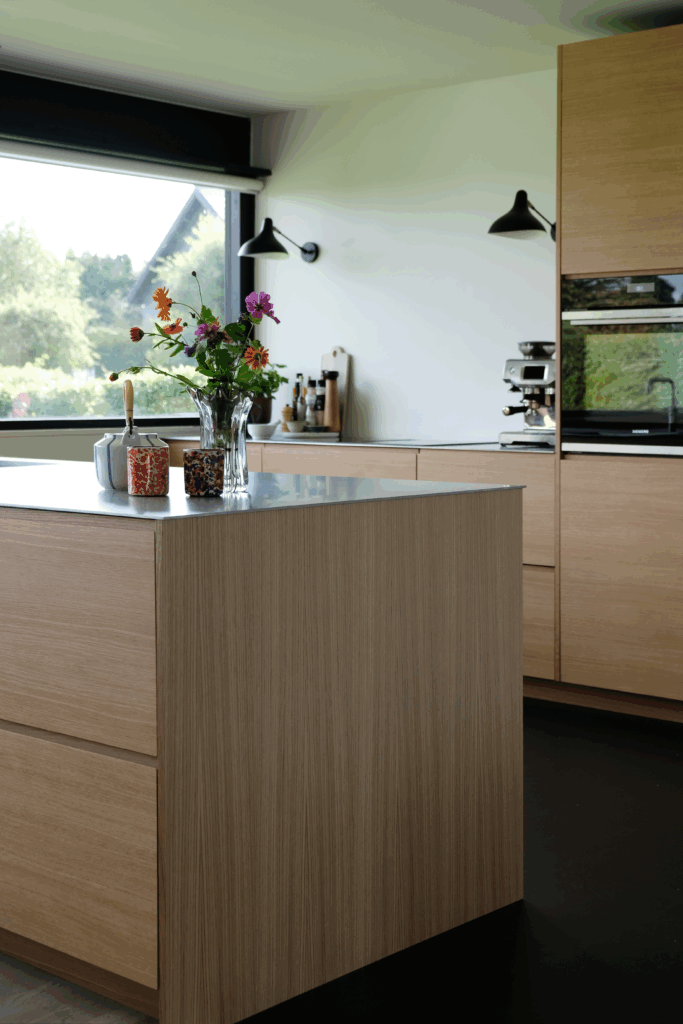 Køkken i egetræ med store vinduer og friske blomster på bordet – inspiration til hvordan man gør et køkken hyggeligt med naturlige materialer og personlig stemning.