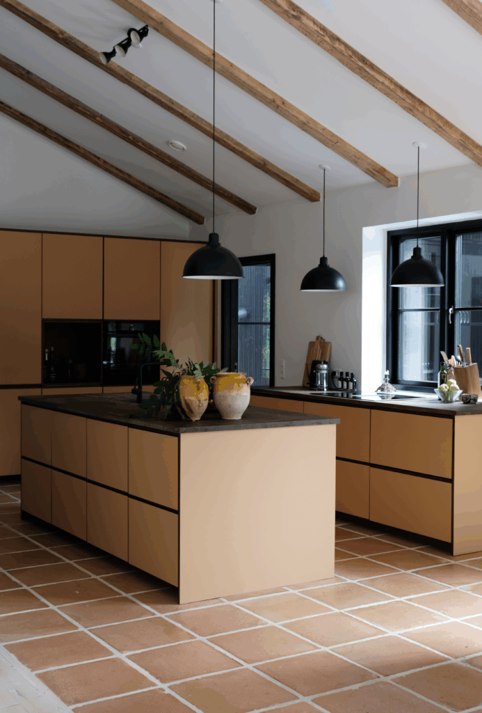 Moderne gult køkken med mørk bordplade, synlige loftsbjælker og store sorte lamper – et varmt og stilrent eksempel på gult køkken inspiration.