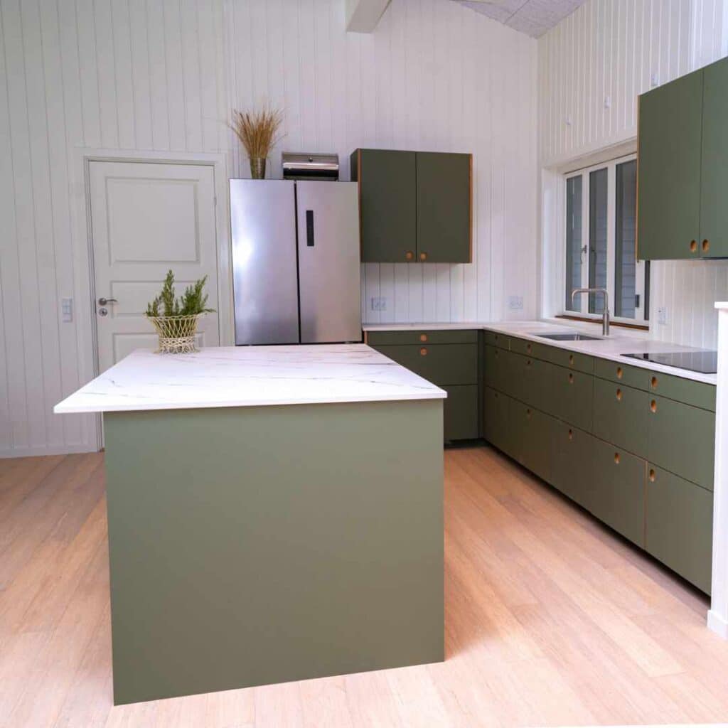 Olivengrøn køkken i sommerhus med køkkenø, lyse vægge og moderne opbevaring.
