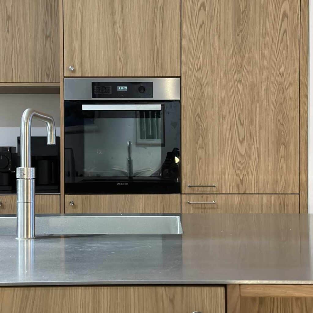 Mørkt køkken med træfronter, stålbordplade og moderne hvidevarer i et stilrent og tidløst design.