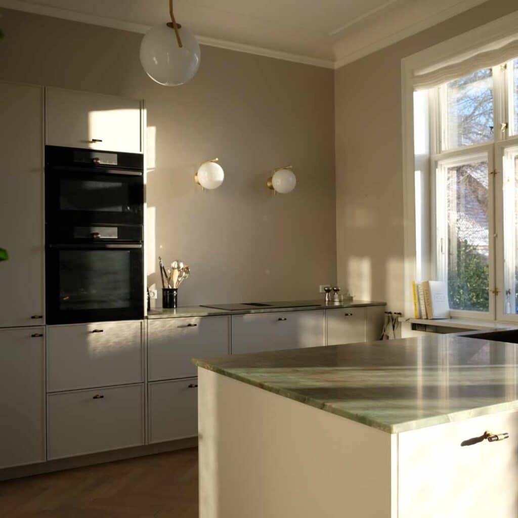 Lyst køkken i varme grønne nuancer med køkkenø, naturstensbordplade og elegante detaljer.