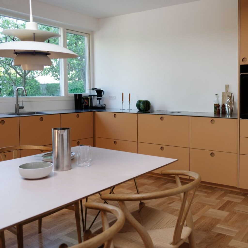 Moderne køkken med varme sandfarvede fronter, sorte bordplader og spiseplads i lyst træ.